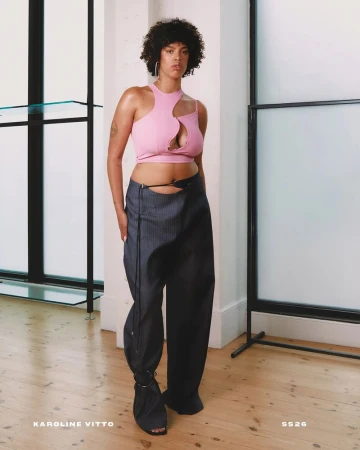 A light-skinned female model poses in a pink halter top with a cutout and dark pinstripe pants in a photo studio with natural light.