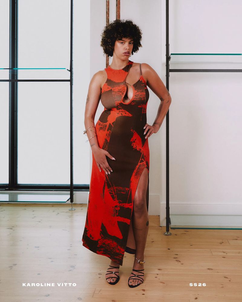 A woman poses in a stylish brown and red abstract-print dress with a thigh-high slit, complemented by strappy heeled sandals indoors.