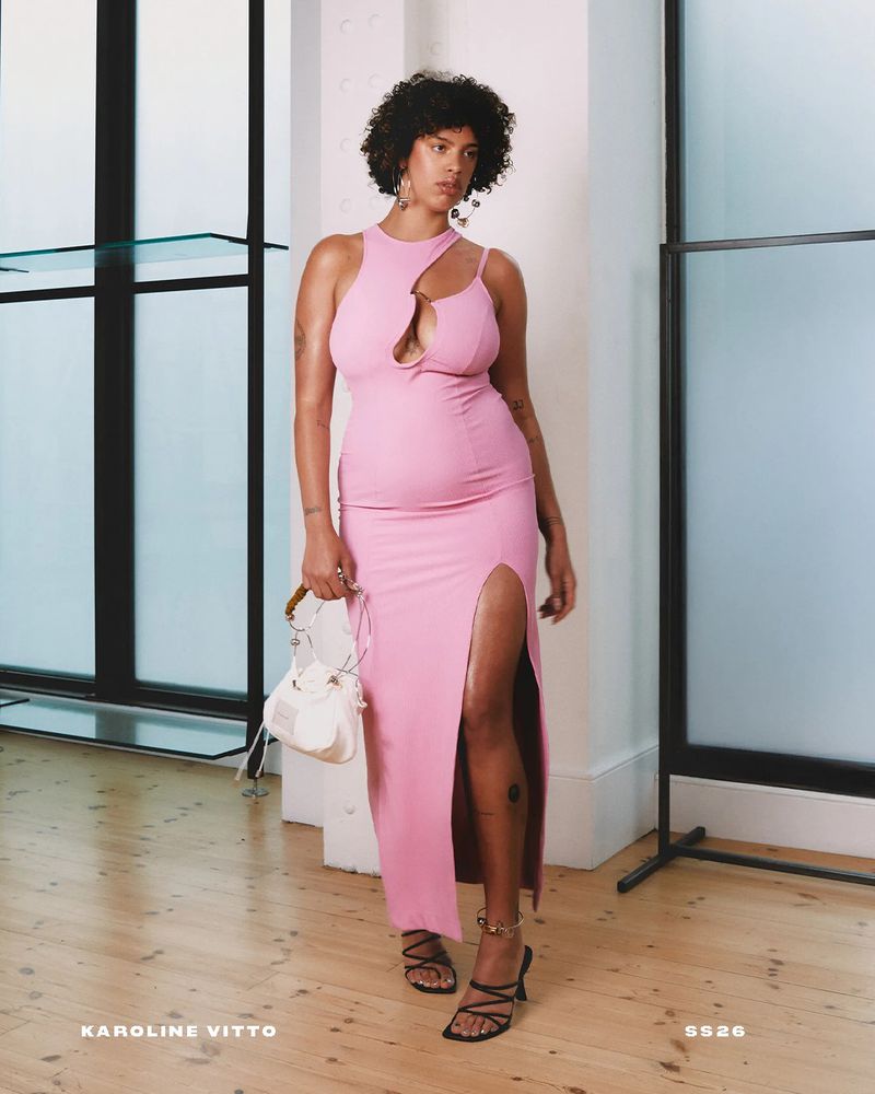 A solo adult female poses in a pink dress with a front slit and strappy sandals in a light-filled indoor studio with minimal props.
