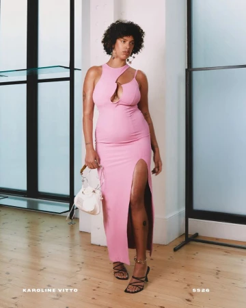A solo adult female poses in a pink dress with a front slit and strappy sandals in a light-filled indoor studio with minimal props.