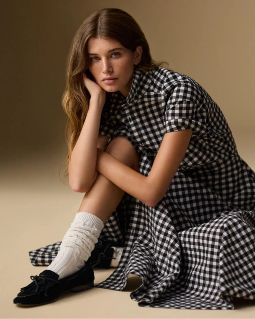 A young woman with long brown hair poses in a black-and-white gingham dress, white socks, and black loafers against a tan background.