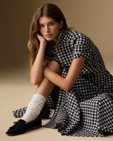 A young woman with long brown hair poses in a black-and-white gingham dress, white socks, and black loafers against a tan background.