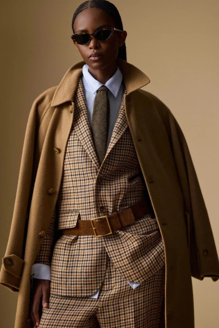 Fashionable woman in a tan plaid suit, brown belt, and oversized tan coat with retro sunglasses, shot in a neutral-toned studio.