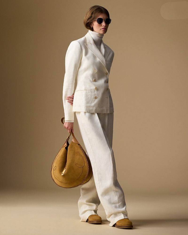 Elegant woman in a white suit and sunglasses carrying a tan suede handbag, posing against a neutral backdrop in a fashion shot.