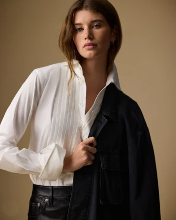 Model wearing a pleated white button-down shirt, black jacket draped over one shoulder, and black pants against a beige studio backdrop.