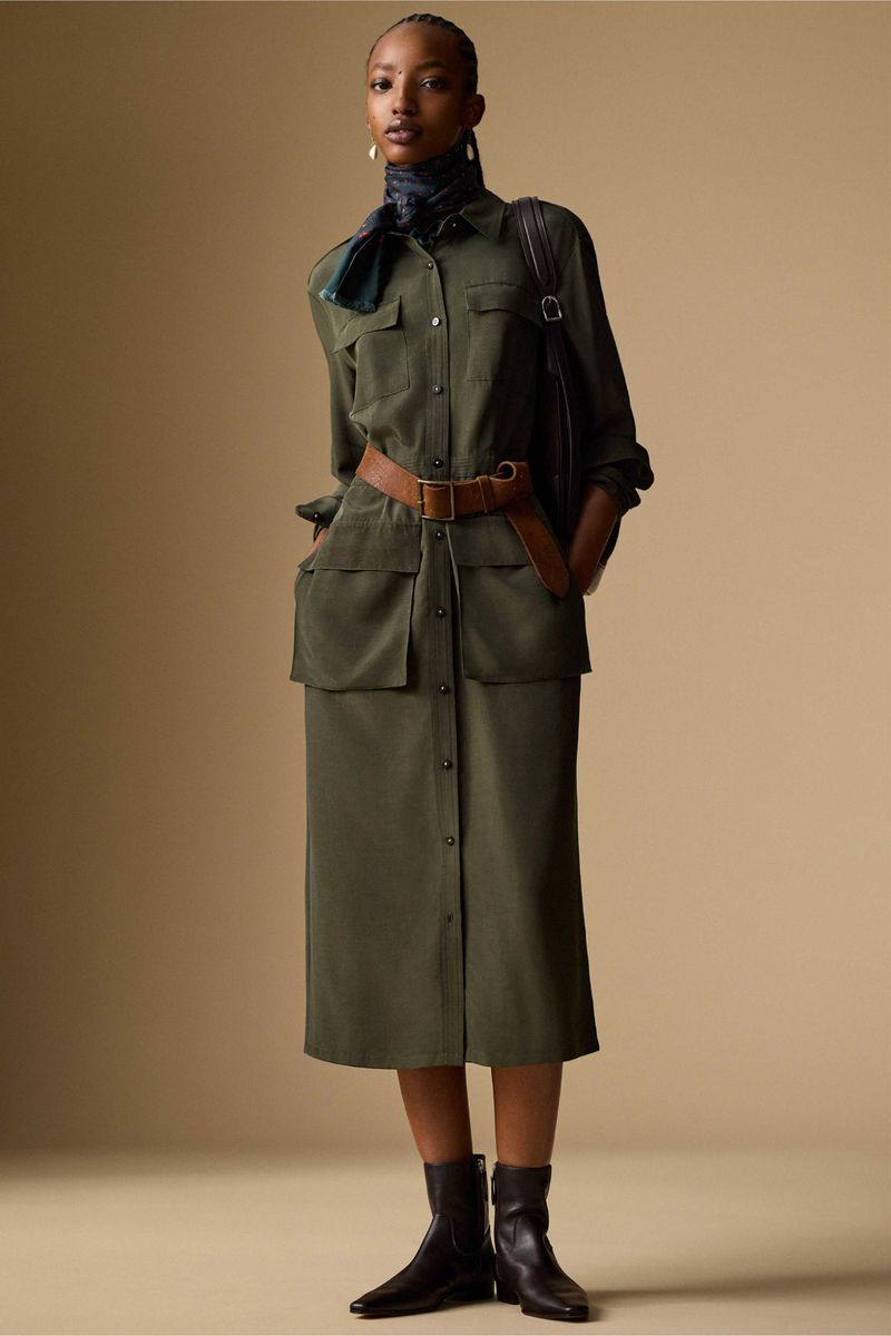 Fashionable woman in an olive green utility dress, brown belt, and black ankle boots posing in a studio with a neutral background.