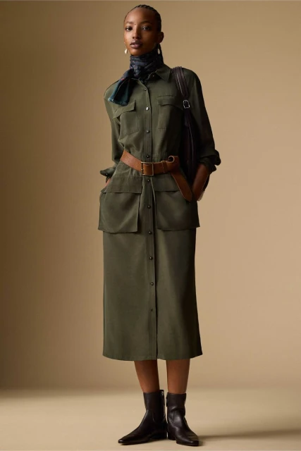 Fashionable woman in an olive green utility dress, brown belt, and black ankle boots posing in a studio with a neutral background.