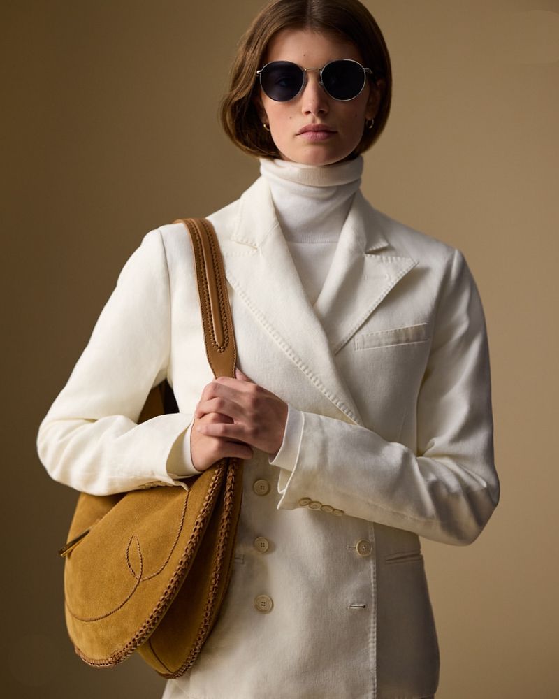Fashionable woman in cream suit and sunglasses carrying a tan suede shoulder bag, posing indoors against a neutral background.