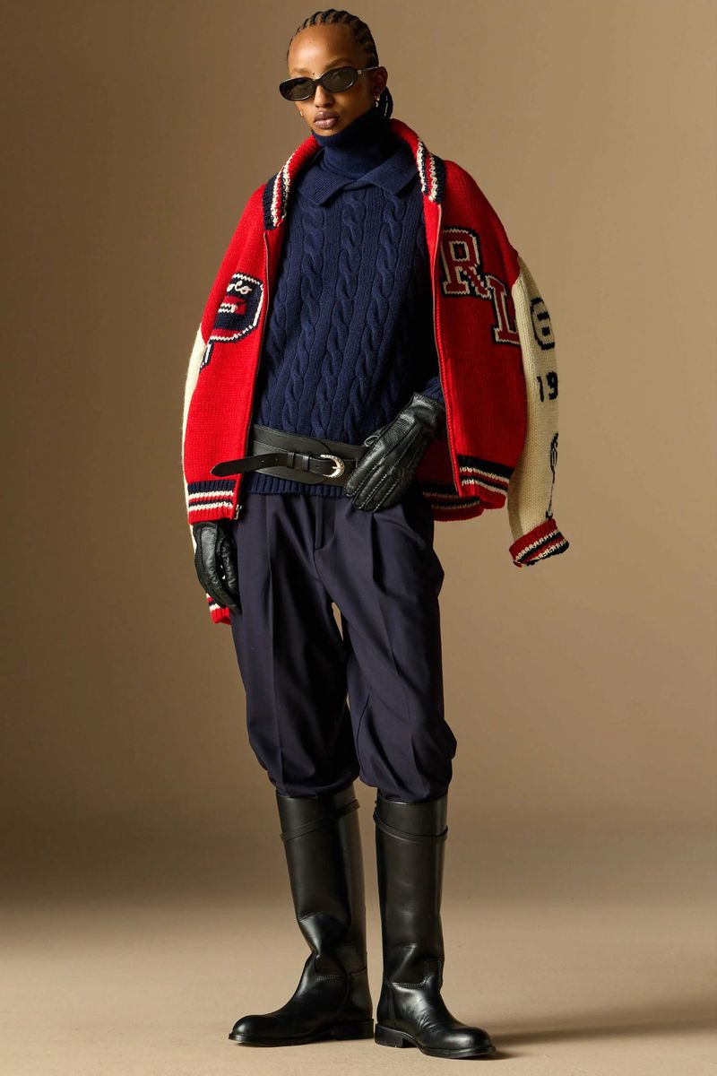 Fashion model wearing a letterman jacket, cable knit sweater, and tall boots in a studio shot for a fashion editorial.
