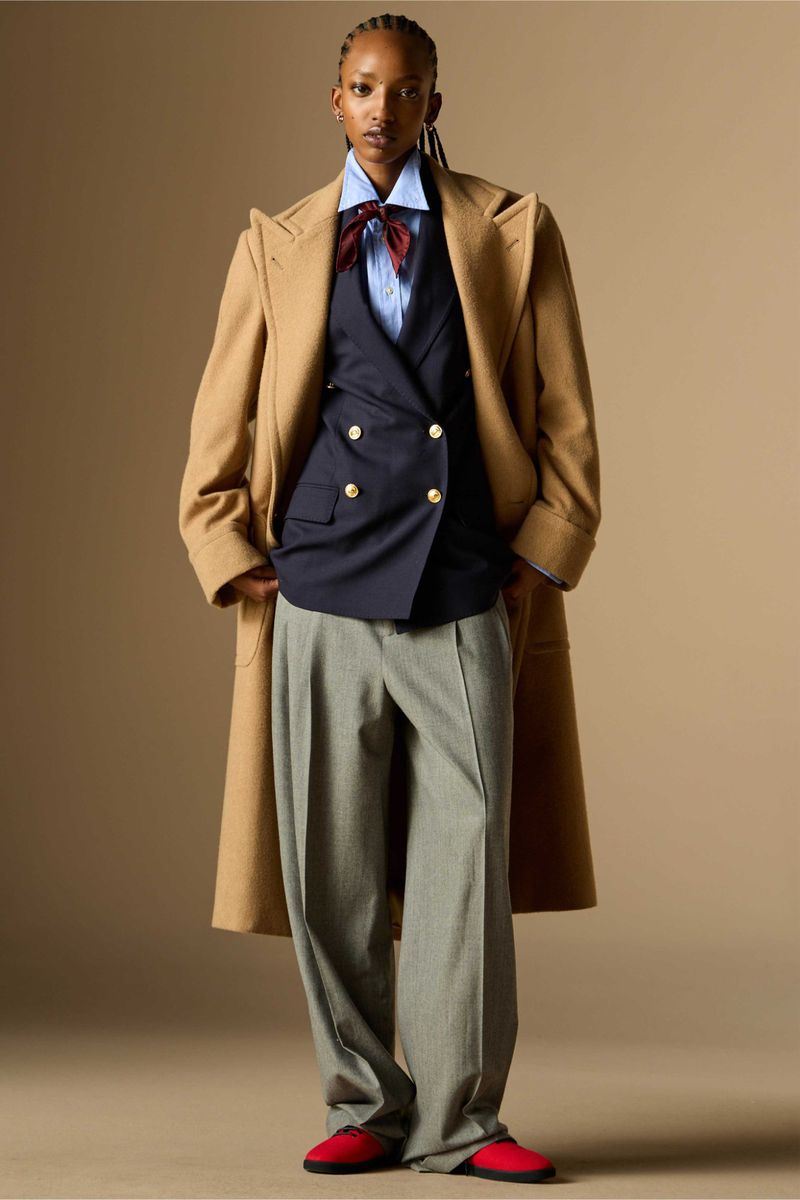 Full-length shot of a Black female fashion model in a beige coat, navy vest, light-blue shirt, bow tie, gray pants, and red shoes against neutral backdrop.