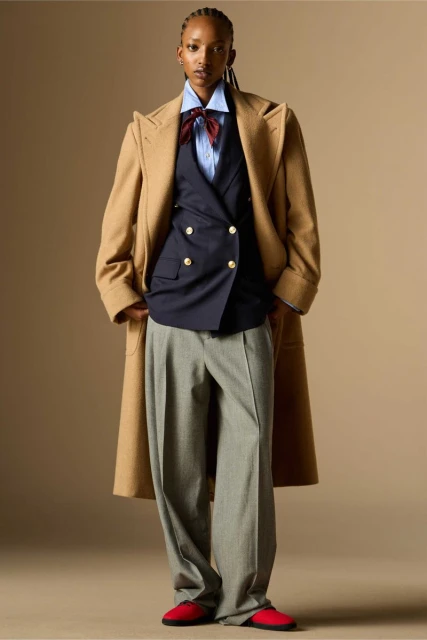 Full-length shot of a Black female fashion model in a beige coat, navy vest, light-blue shirt, bow tie, gray pants, and red shoes against neutral backdrop.