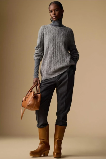 Fashion shot of an adult woman wearing a grey sweater, dark pants, and brown boots, holding a brown purse against a tan background.