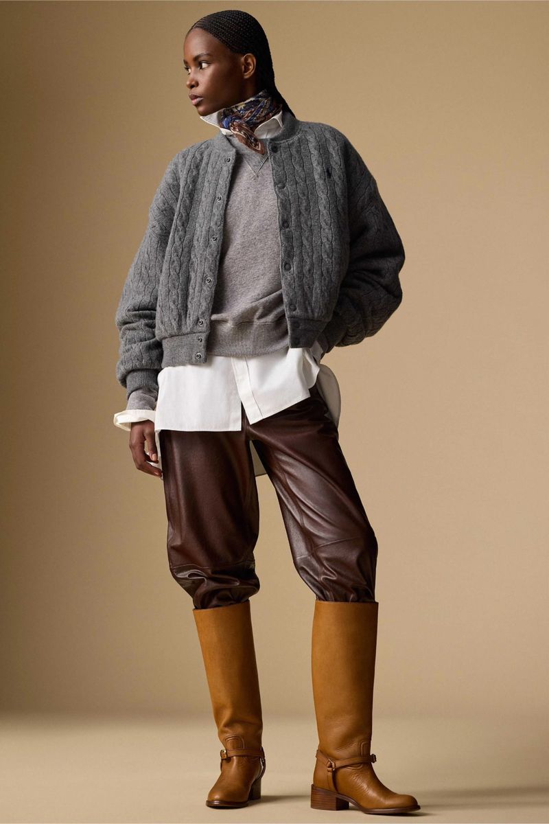Fashion model poses in brown leather jodhpurs and riding boots, wearing a grey sweater and cardigan against a tan backdrop.