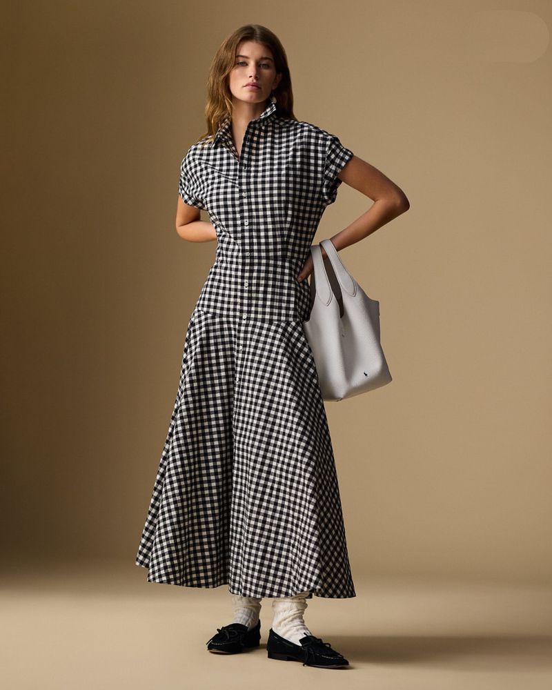 A woman stands in a photo studio wearing a black-and-white checkered dress, white socks and black shoes, carrying a gray tote bag.