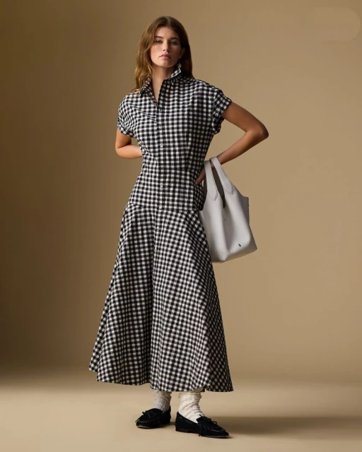 A woman stands in a photo studio wearing a black-and-white checkered dress, white socks and black shoes, carrying a gray tote bag.