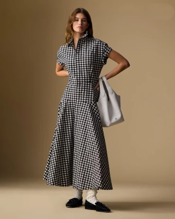 A woman stands in a photo studio wearing a black-and-white checkered dress, white socks and black shoes, carrying a gray tote bag.