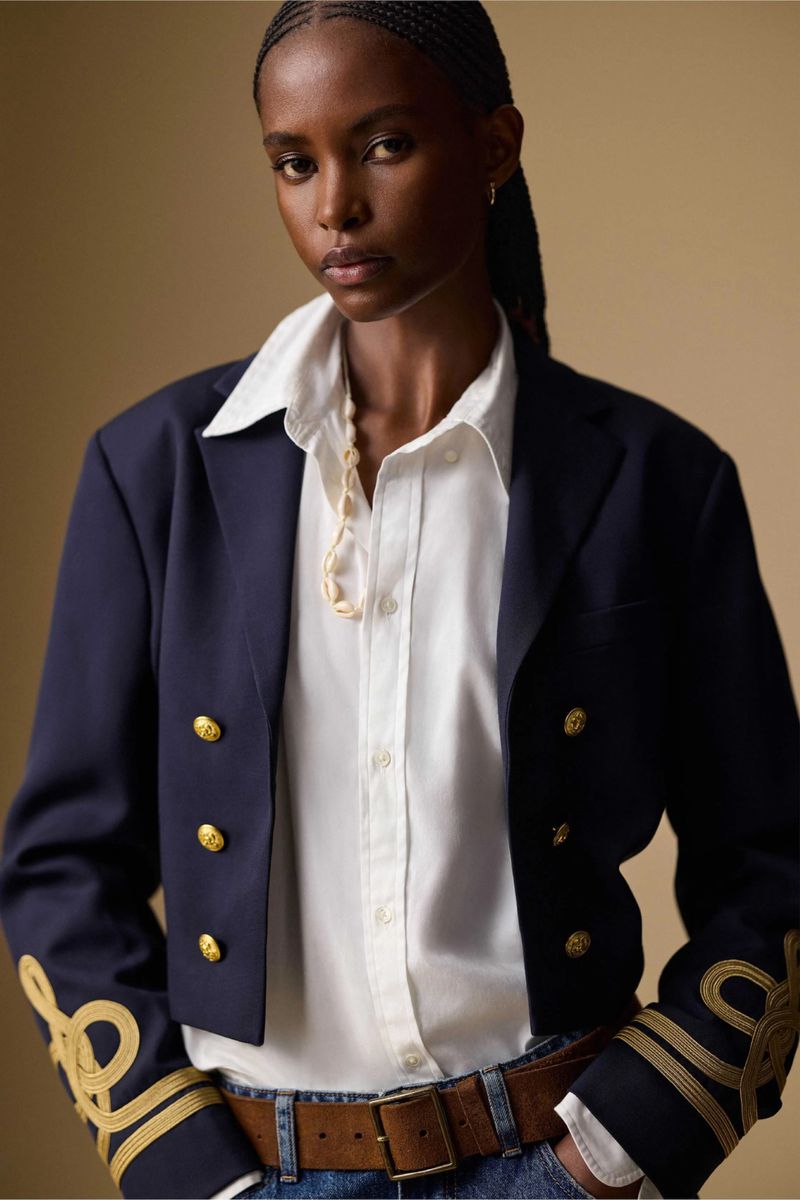 Dark-skinned woman in a navy blazer with gold accents, white button-down shirt, jeans with brown belt poses in a studio setting.