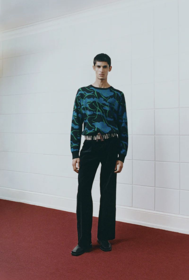 Full-length shot of a young adult male in a patterned sweater, black pants, and leather shoes, standing on a red carpet in a corridor.