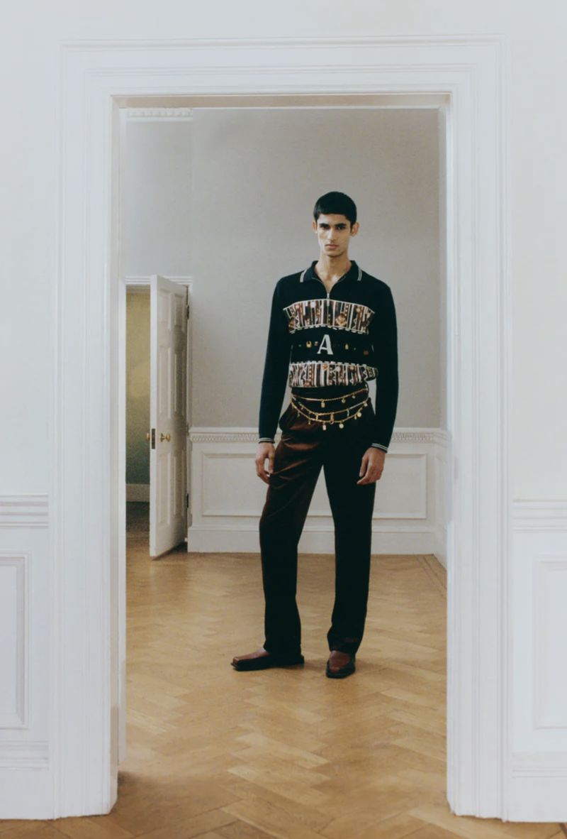 Model wearing a black graphic zip-up knit sweater, dark trousers, and a gold chain belt framed in an interior doorway.