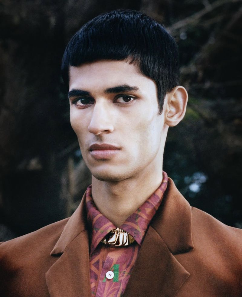 Portrait of an adult male model with dark hair and brown eyes wearing a brown suit jacket and patterned shirt, posing outdoors.