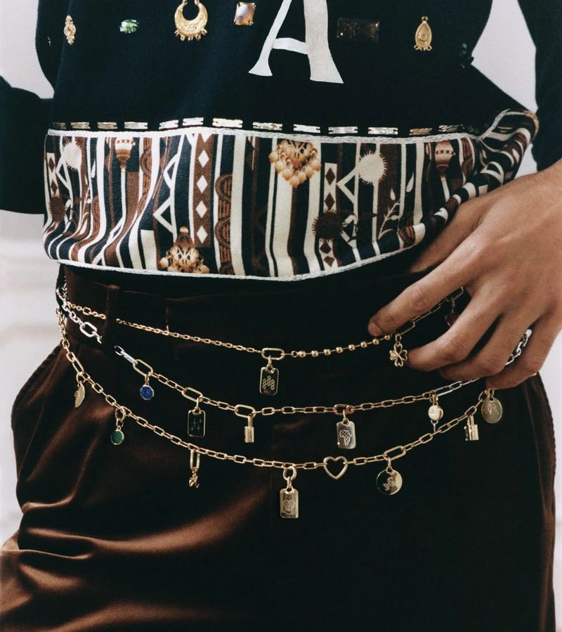 Close-up of a person wearing a black sweater and brown trousers, accessorized with three gold chain belts featuring charm details.
