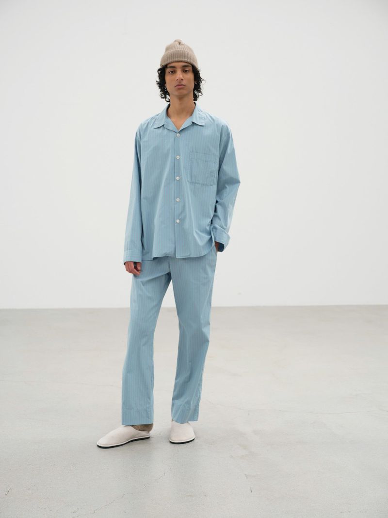 A young adult male stands in a studio wearing a light blue pajama set and a beanie, looking directly at the camera.