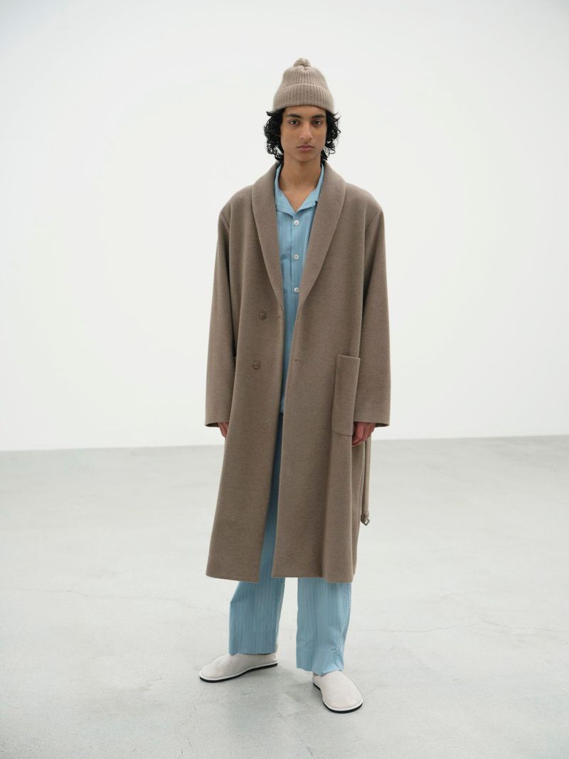 Man in tan coat and beanie, light blue pajamas, and white shoes stands in a white room looking at the camera in a fashion shot.