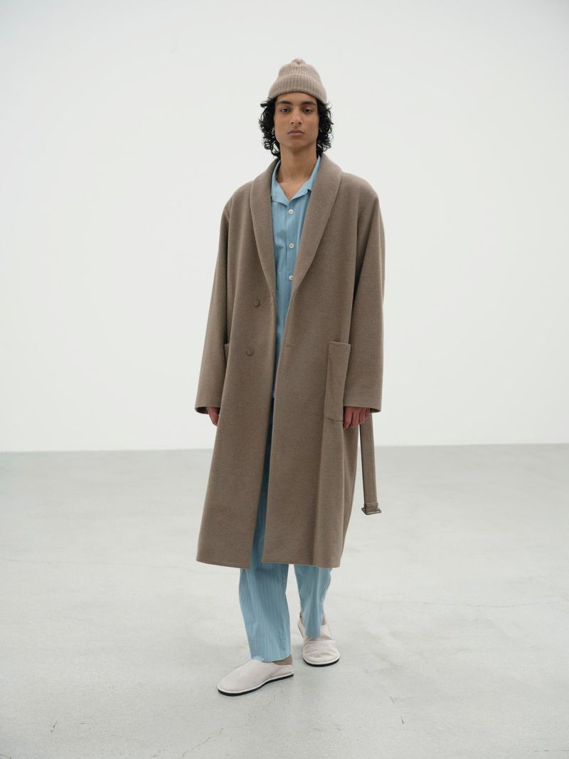 A person standing indoors, wearing a long brown coat, light blue pajamas, a beige beanie, and white shoes, on a white studio backdrop.