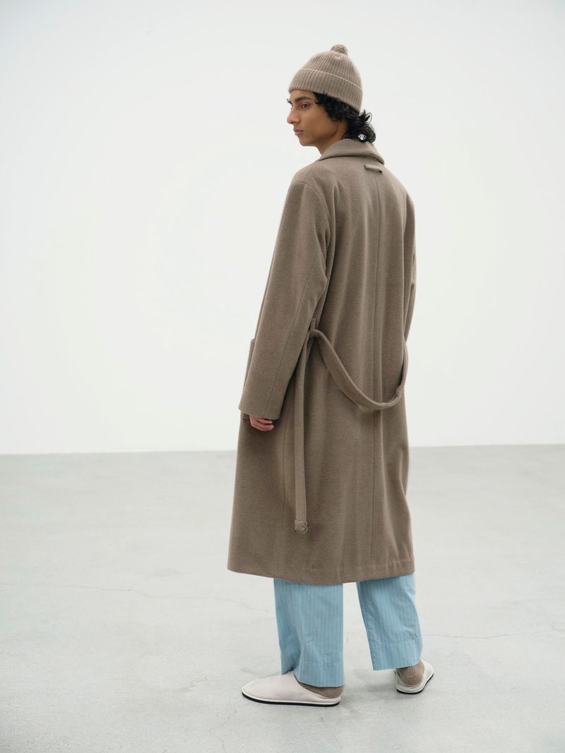 Max Everist wearing a taupe belted overcoat, blue trousers, and a beanie against a white backdrop for Auralee F/W 25.