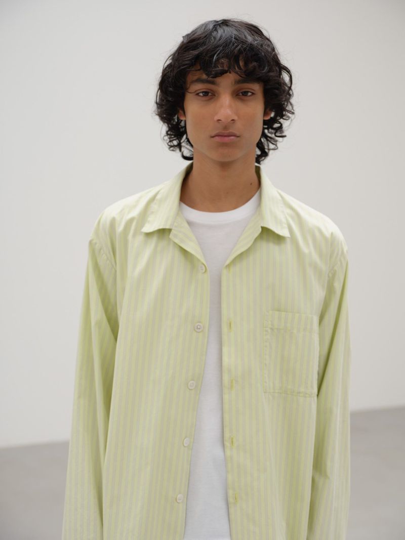 A young adult with curly hair stands indoors wearing a striped light green shirt and white T-shirt against a bright white backdrop.