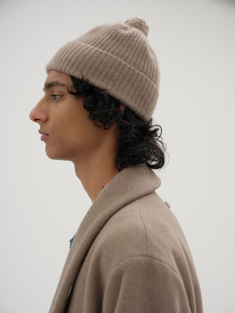 Profile view of an adult with dark curly hair wearing a beige beanie hat with a pompom and a beige coat against a white backdrop.