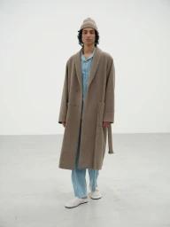 Model Max Everist wearing a taupe wool overcoat, knit beanie, and light blue set against a minimal neutral studio backdrop.