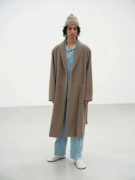 Model wearing a long taupe wool overcoat, light blue matching shirt and pants, and a beige knit beanie in a bright studio.
