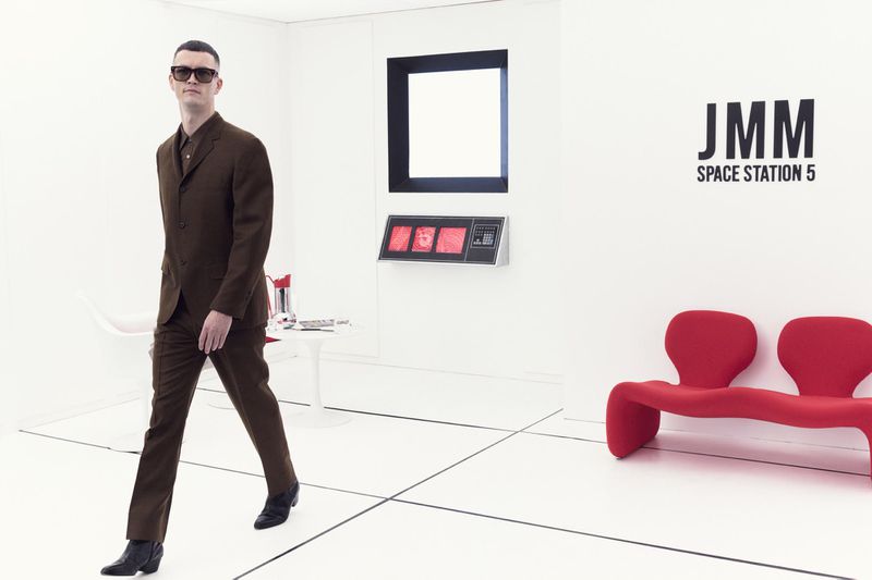 A man in a brown suit walks across a white tiled floor in a stark white room with minimalist furniture and the text 'JMM Space Station 5'.