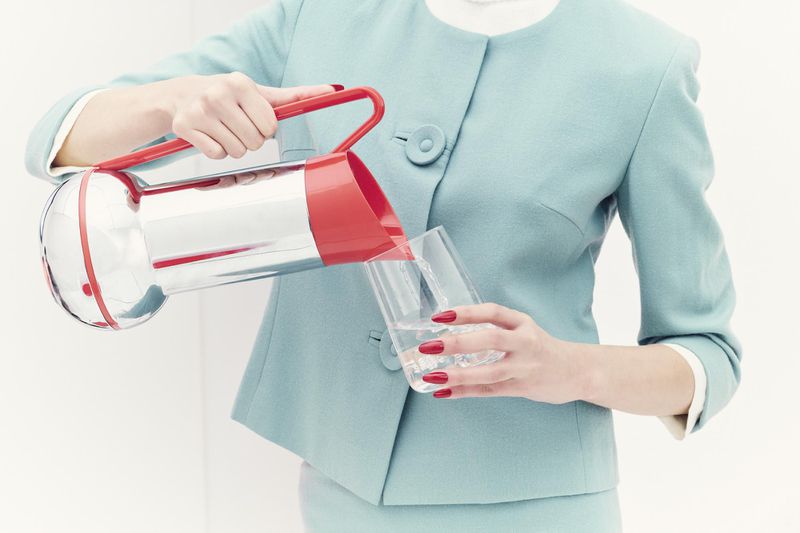 Woman in a teal skirt suit pours water from a red and silver pitcher into a glass against a white background in a studio shot.