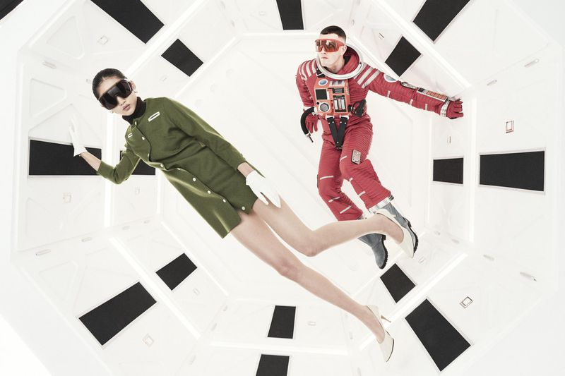 A female model in a green dress and a male model in an astronaut suit pose in a bright white, octagonally shaped futuristic interior.
