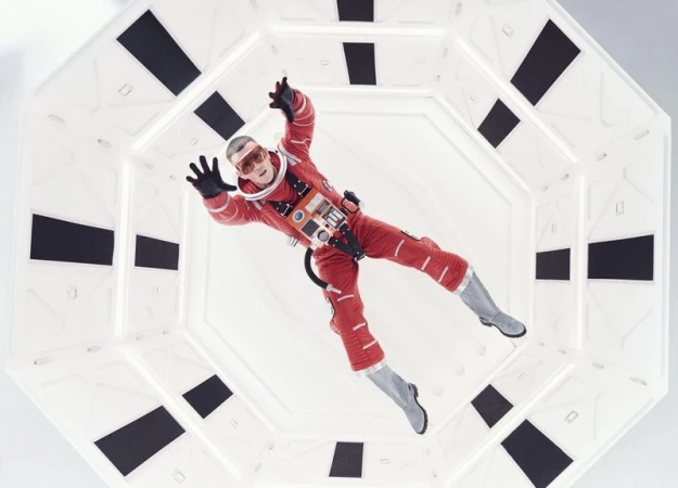 Man in a red spacesuit floats inside a white, octagonal-shaped hallway with black rectangular accents, captured in a full shot.