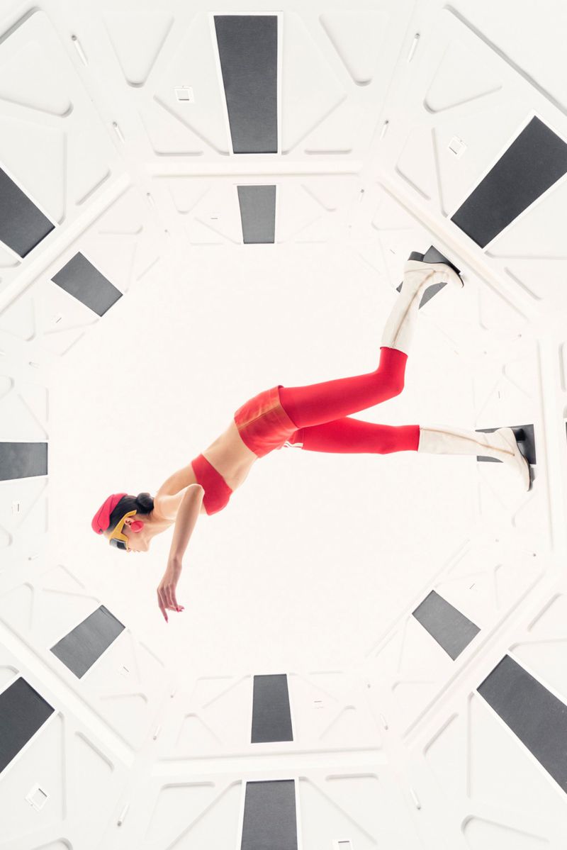 Woman in red outfit poses inside white geometric tunnel-like space, viewed from below, creating a sense of perspective and modern style.