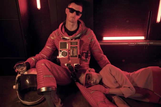 A man in a red spacesuit sits with a woman lying down and a glass of liquor in a red-lit interior scene with an astronaut helmet nearby.