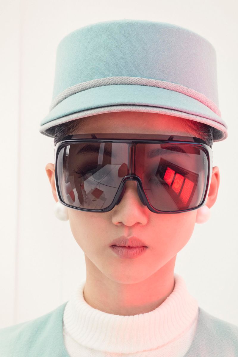Close-up of a person wearing a blue hat and large sunglasses, looking directly at the camera with a neutral expression in a white studio.