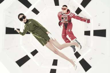 Two models wearing futuristic visor sunglasses appear weightless inside a bright white retro spaceship corridor.
