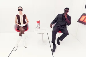 Two models in retro-futuristic styling wearing Jacques Marie Mage sunglasses seated in a bright white minimalist room.