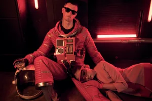 Man in a red astronaut suit holding a glass with a woman resting beside him in a dimly lit red-toned interior