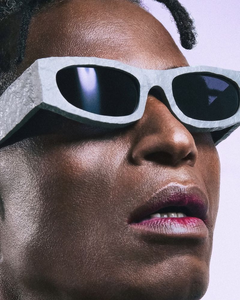 Close-up of a person wearing large grey sunglasses with an angular design, magenta lipstick, against a light purple backdrop.
