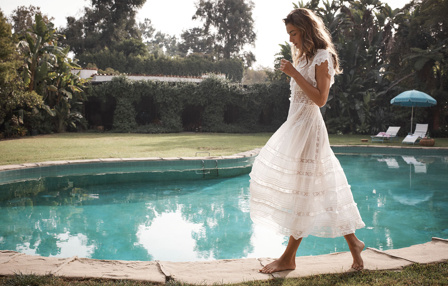 A woman in a white dress walks along a stone edge by a pool in a sunlit outdoor setting with lush greenery and an umbrella in the background.