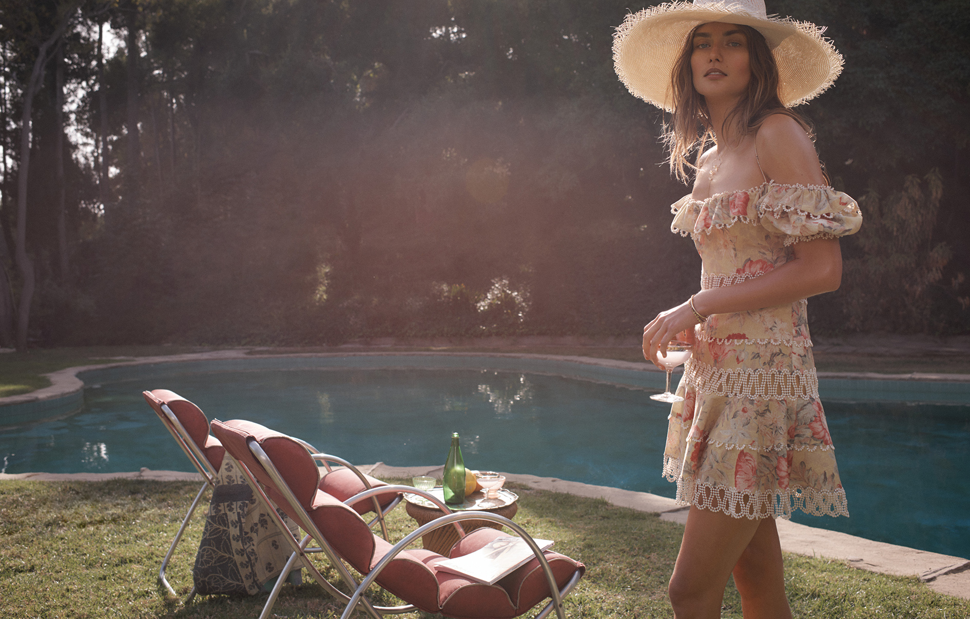 Brunette woman in floral mini dress and straw hat, outdoors on a sunny day, holding a drink by the pool, with lounge chairs.