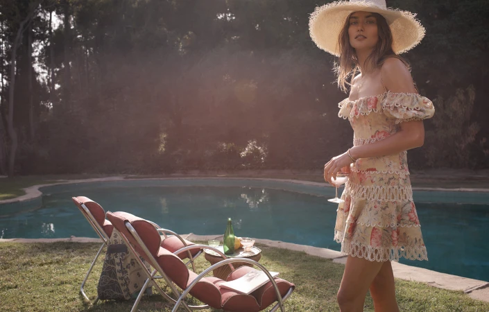 Brunette woman in floral mini dress and straw hat, outdoors on a sunny day, holding a drink by the pool, with lounge chairs.