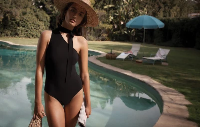 Model in black swimsuit and straw hat standing by a swimming pool holding a patterned clutch.