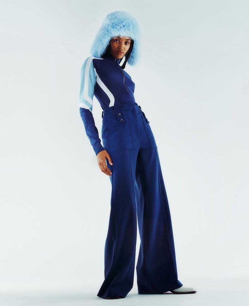Fashion model in a blue jumpsuit with a furry blue hat, posing in a studio setting with a white background, looking at the camera.
