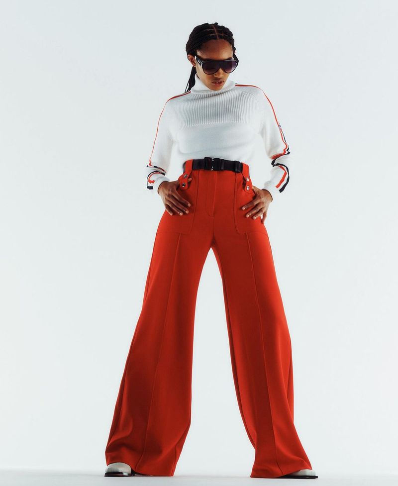 A fashionable woman poses in a white turtleneck with red accents and high-waisted, wide-legged red pants against a bright white backdrop.